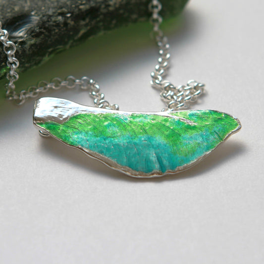 Pendant with the shape of a maple key approximately 44mm long by 16mm wide made of pure silver with transparent green and blue glass enamel accent colour. Sterling silver chain 18" long. The maple key hangs horizontally with the chain attached to the back side of the pendant.